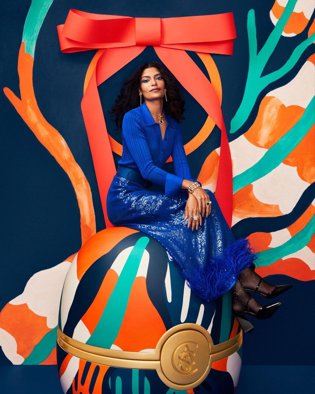 Elegant woman in blue sits atop a large, patterned ornament with a red bow, set against a deep blue backdrop with abstract decorations.