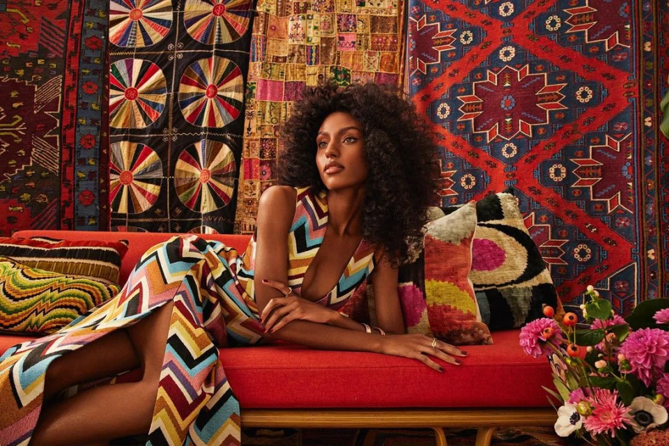 A woman with dark hair and a colorful patterned dress poses on a red couch in front of patterned textiles and floral arrangement.