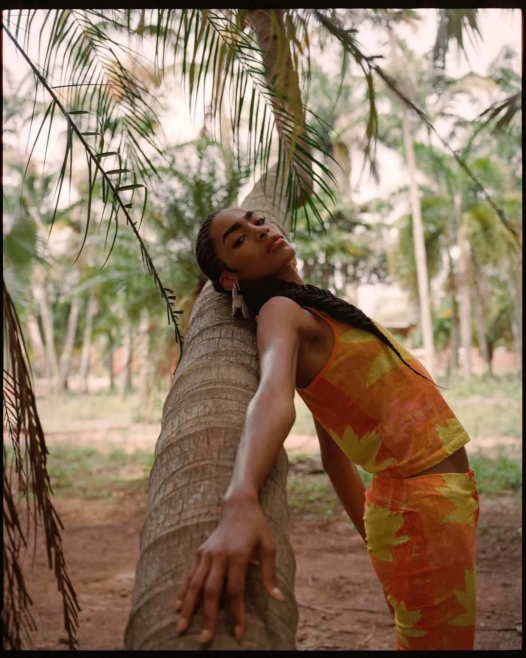 Young woman in a patterned outfit leans against a palm tree in a tropical setting with many palm trees in the background during daytime.