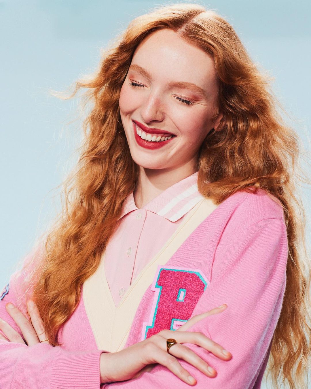 Smiling red-haired woman in a pink cardigan with arms crossed against a light blue background. Close-up portrait.