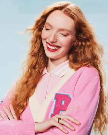 Smiling red-haired woman in a pink cardigan with arms crossed against a light blue background. Close-up portrait.
