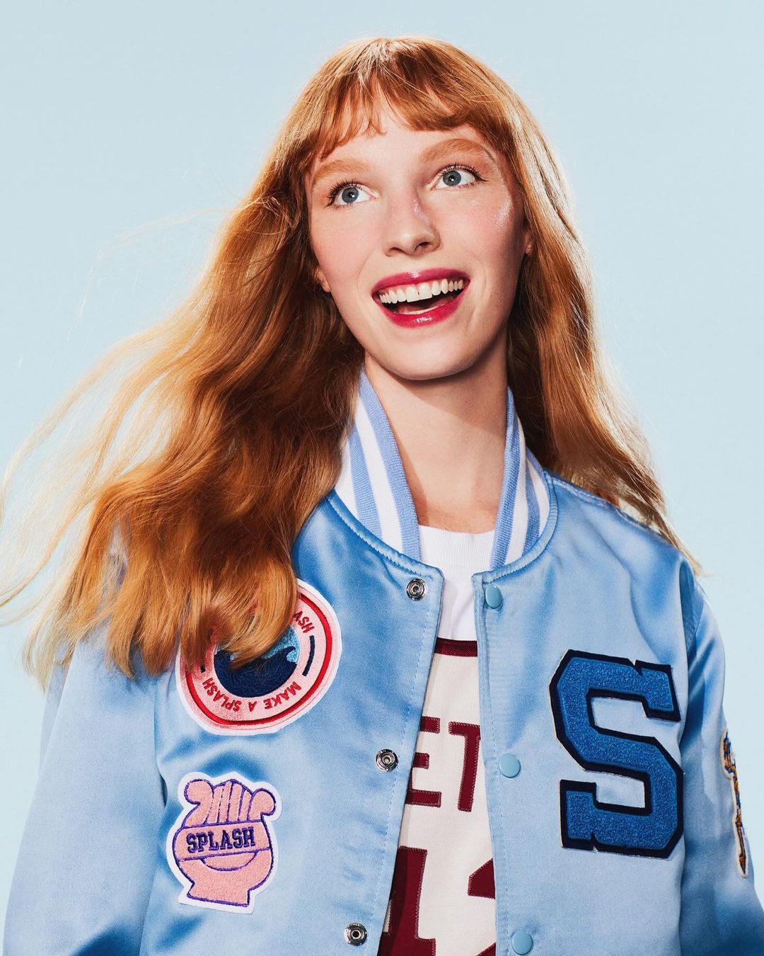 Portrait of a red-haired woman in a light blue letterman jacket smiling and looking up against a solid light blue background.