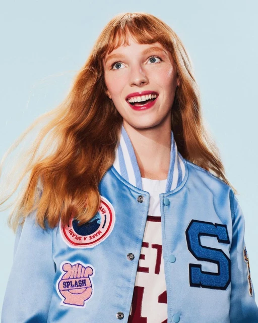 Portrait of a red-haired woman in a light blue letterman jacket smiling and looking up against a solid light blue background.