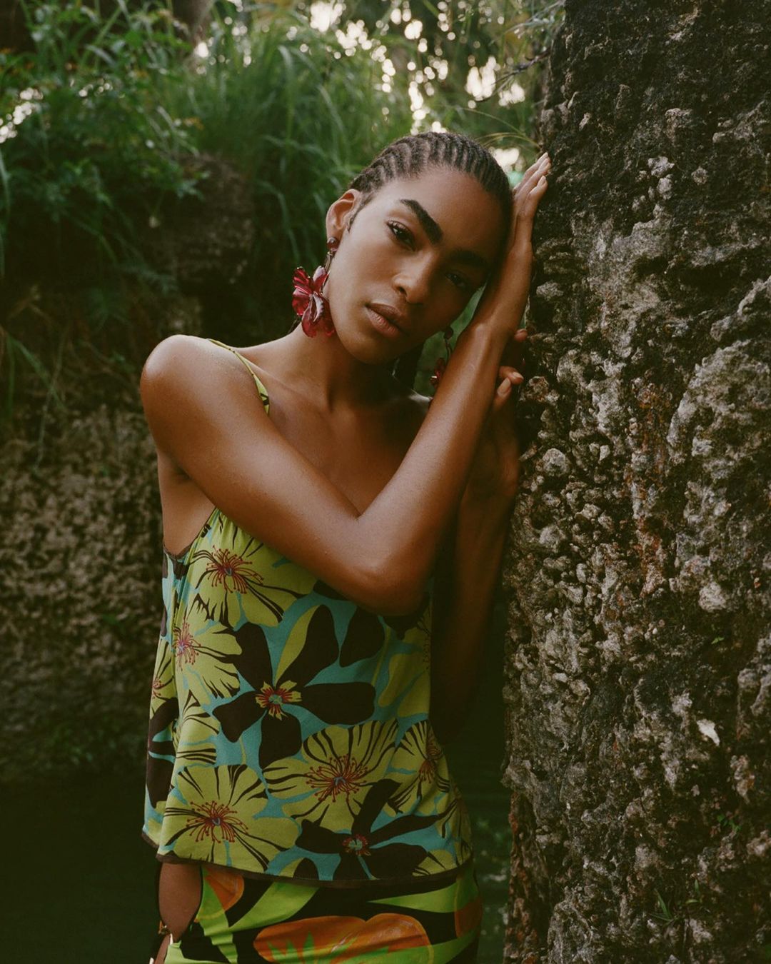 A fashionable young adult female model poses leaning against a textured stone surface wearing a floral print top with tropical background.