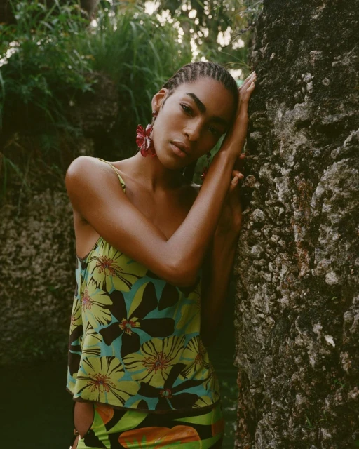 A fashionable young adult female model poses leaning against a textured stone surface wearing a floral print top with tropical background.