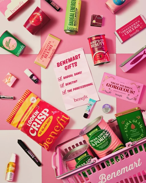 Benefit brand beauty products displayed in a pink shopping basket, arranged on a pink-and-white checkered surface in a studio setting.