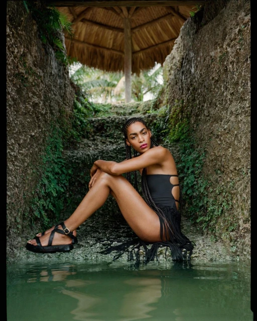 A woman in a black swimsuit with fringe detail sits in a rocky grotto with water; a straw-roofed structure is visible in the background.