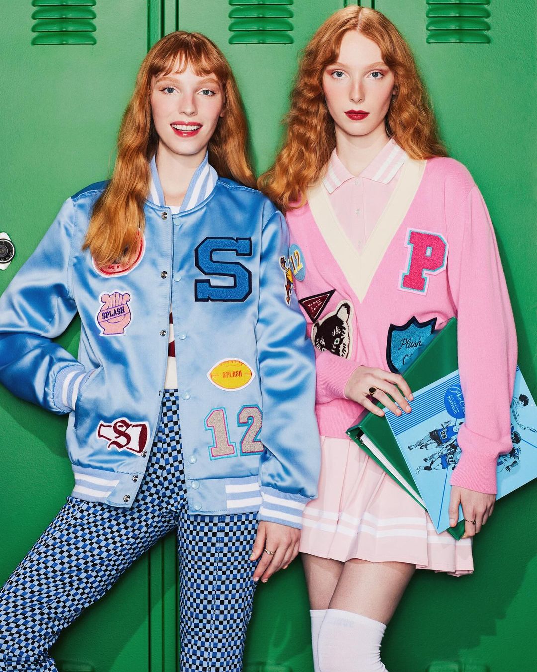 Two red-haired women in retro letterman jackets and preppy outfits pose in front of a green locker-lined wall for a fashion editorial.