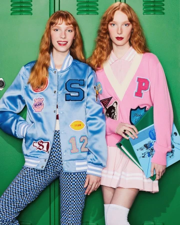 Two red-haired women in retro letterman jackets and preppy outfits pose in front of a green locker-lined wall for a fashion editorial.