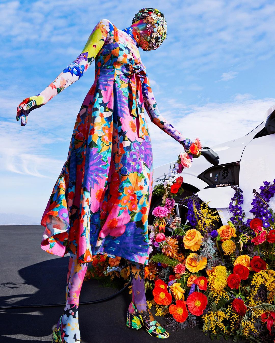 Model in vibrant floral dress stands next to a flower-covered electric car against a blue sky, merging fashion with automotive.