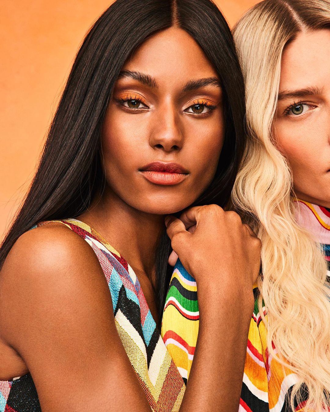 Close-up studio shot of two women with orange eyeshadow and patterned tops against an orange backdrop, beauty and fashion focused.