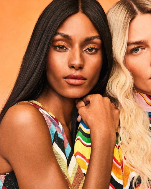Close-up studio shot of two women with orange eyeshadow and patterned tops against an orange backdrop, beauty and fashion focused.
