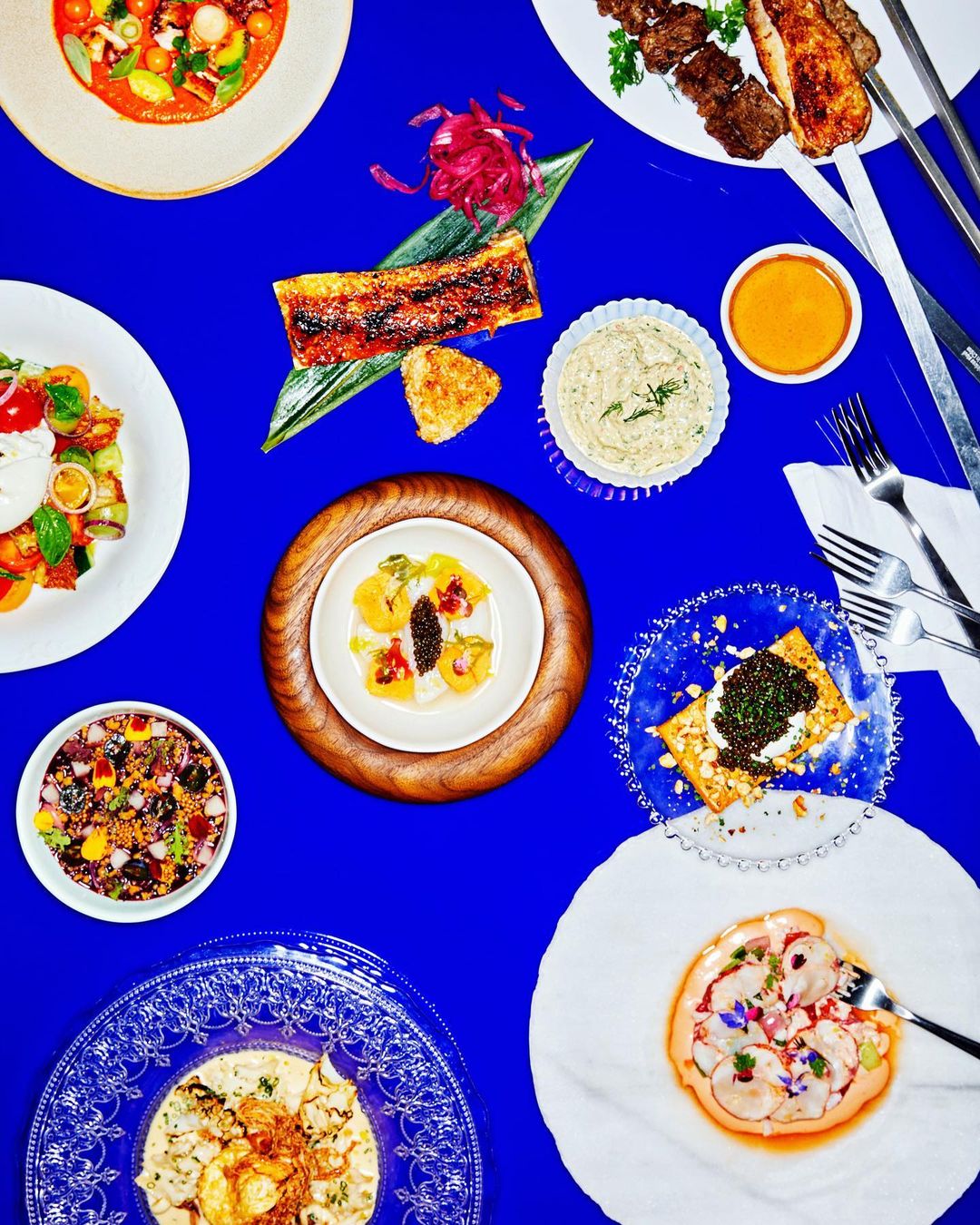Overhead shot of an arrangement of various plated food dishes on a vibrant blue surface, including seafood, meats, and salads.