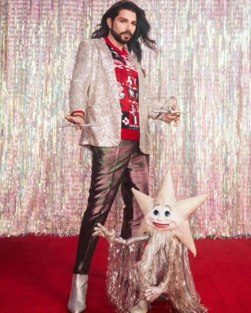 Full shot of a male fashion model in a sequined blazer and star puppet in front of a silver fringe curtain on a red carpet.