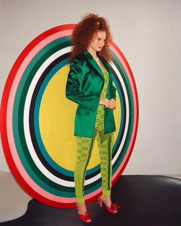 Woman with voluminous red hair wearing a green satin blazer and patterned green leggings standing before a colourful circular art backdrop.