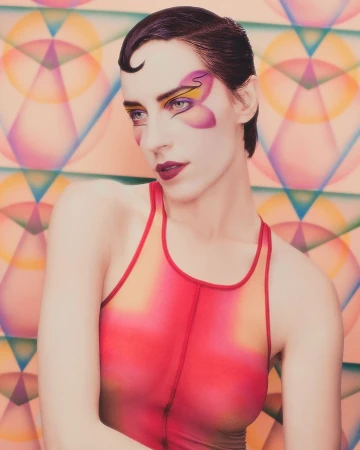 Close-up portrait of a woman with butterfly face art and short dark hair wearing a pink and yellow top against a geometric background