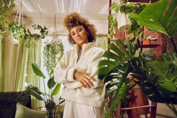Woman with curly blonde hair wearing a white fleece zip jacket standing indoors among hanging plants and large green monstera leaves.