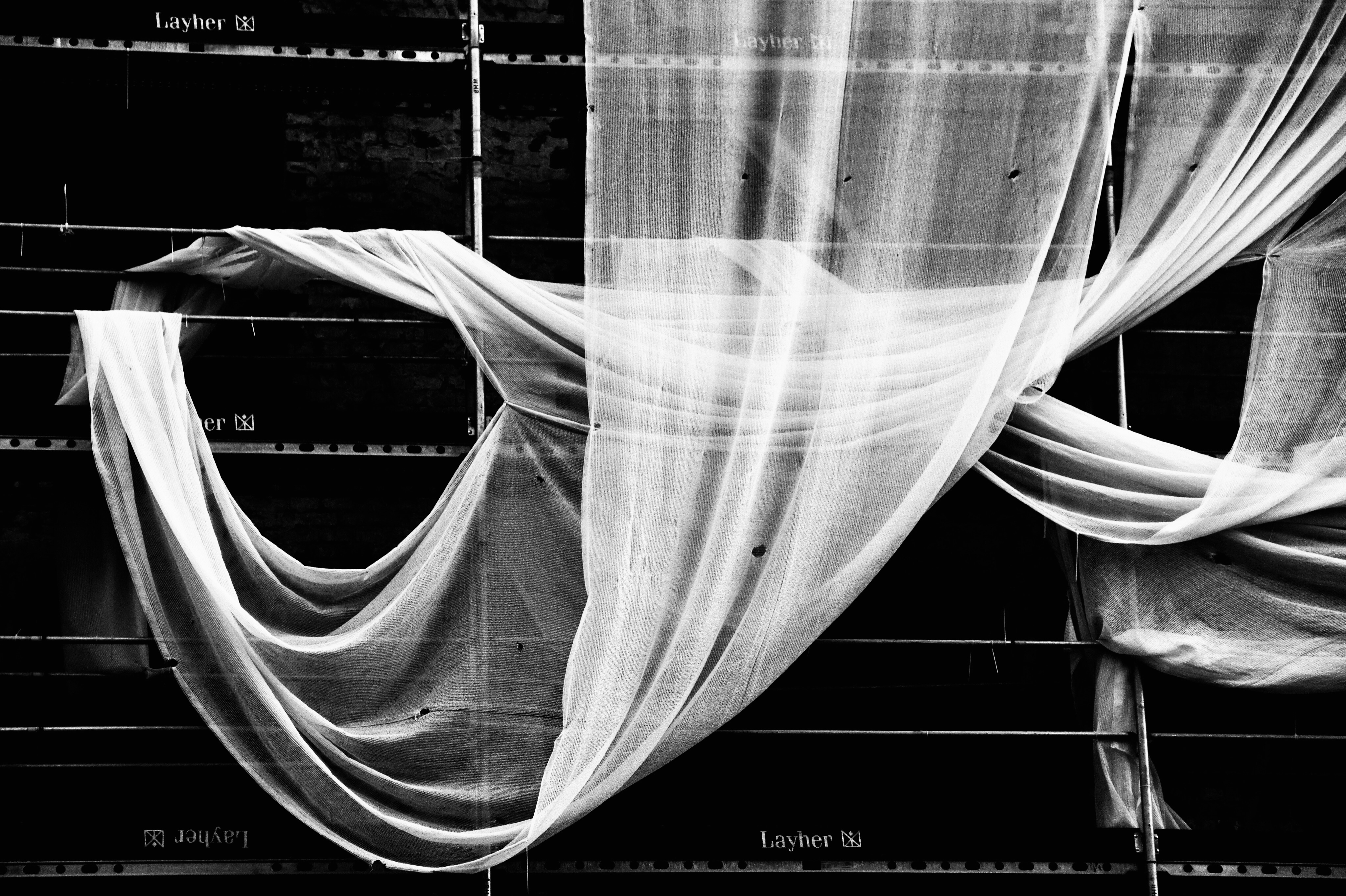 Black and white photo of draped white fabric over scaffolding, creating abstract shapes and patterns with industrial structure visible.