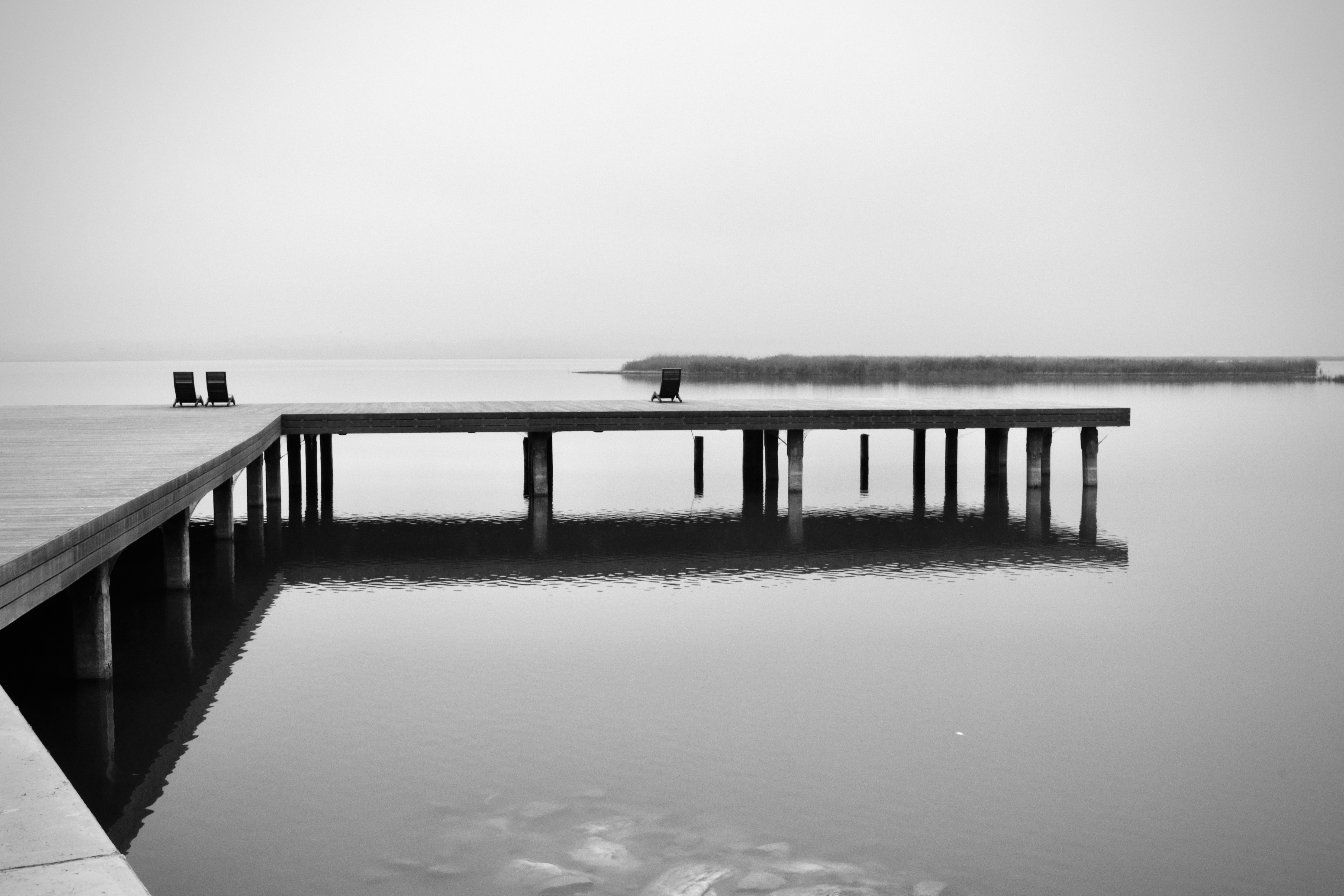 Monochrome landscape featuring a wooden pier extending into a serene body of water, two chairs on the pier, and distant shoreline.