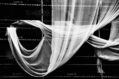 Monochrome architectural photograph of sheer white protective fabric draped across dark metal scaffolding.