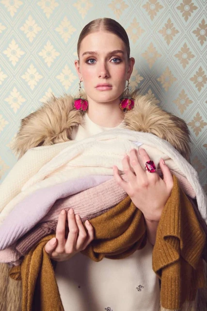 Portrait of a young woman indoors wearing fur-trimmed coat holding a pile of folded sweaters, with bright pink earrings and a pink ring.
