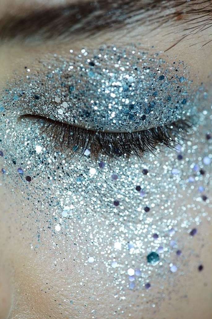 Close-up of a person's eye adorned with shimmering blue glitter makeup. The eye is partially closed, emphasizing the makeup.