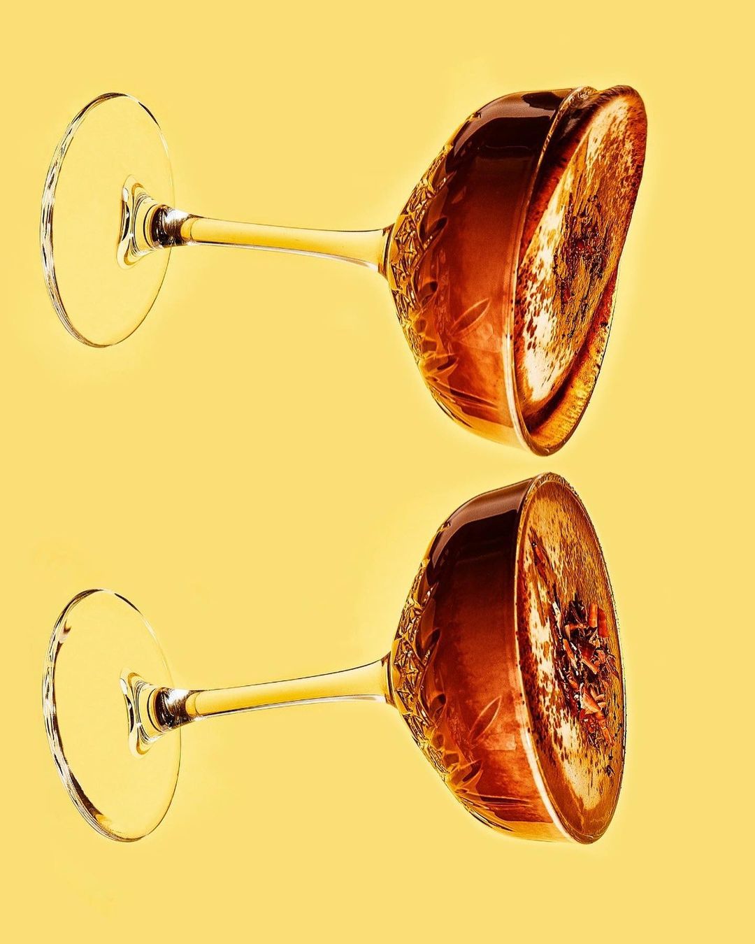 Two wine glasses with ornate designs are presented on a yellow backdrop, showcasing their elegant form and the play of light on glass.
