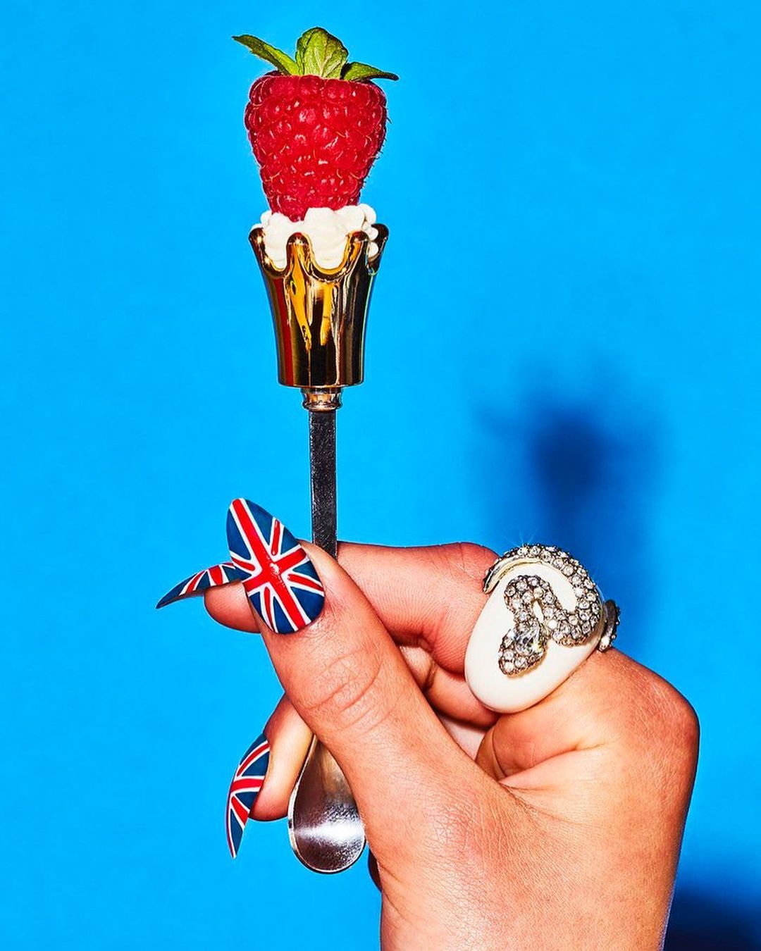 Hand holding a spoon with a raspberry on top against a blue background, showing fashion fingernails with the Union Jack design.