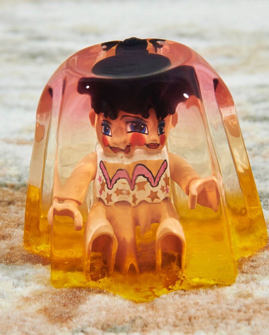 Artistic still life of a three-faced vintage-style toy figure encapsulated in translucent orange resin against a mottled neutral background.