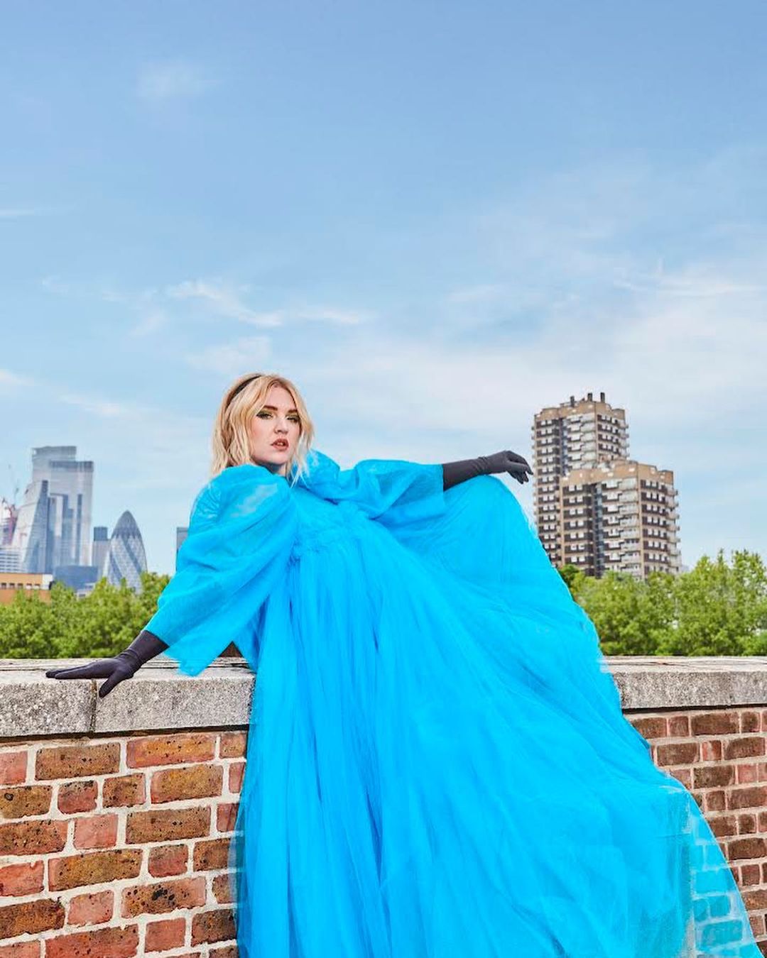 Woman in a blue tulle dress poses on a brick wall, with city buildings in the background under a bright sky, fashion photography.