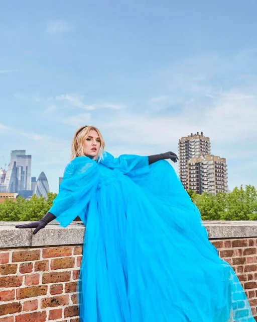 Woman in a blue tulle dress poses on a brick wall, with city buildings in the background under a bright sky, fashion photography.