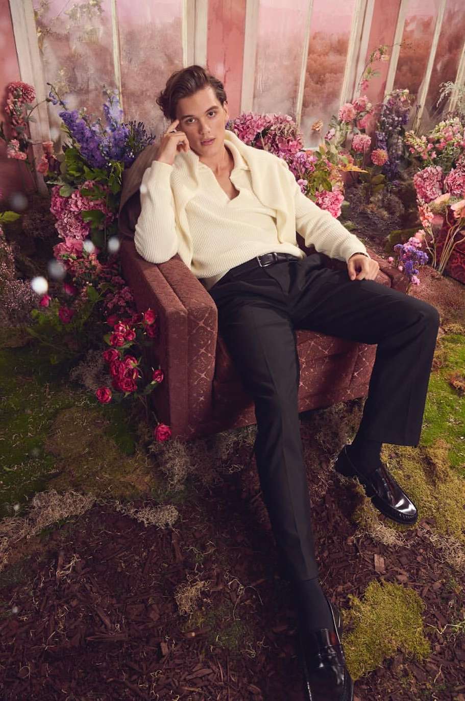Young adult man in cream sweater and black pants sits in a chair surrounded by colorful flowers indoors with a contemplative facial expression.