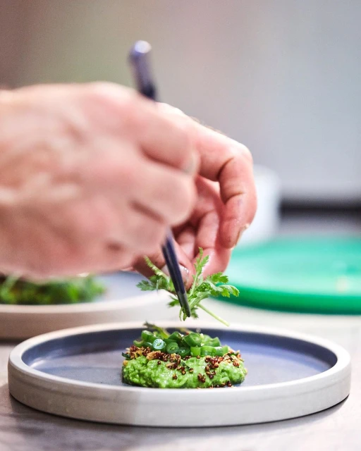 Chef plates food on blue-rimmed white plate. Close-up of greens with herbs and spices with chef's hands using chopsticks.