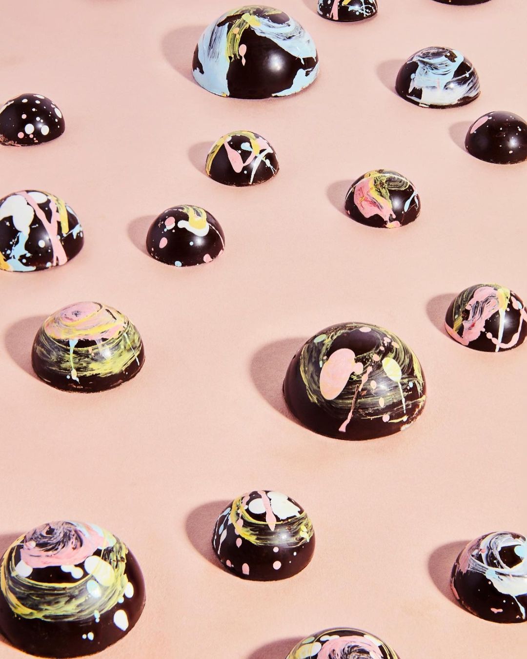 Overhead view of arranged dark chocolate truffles with bright paint splatters on a light pink background. Food and beverage photography.