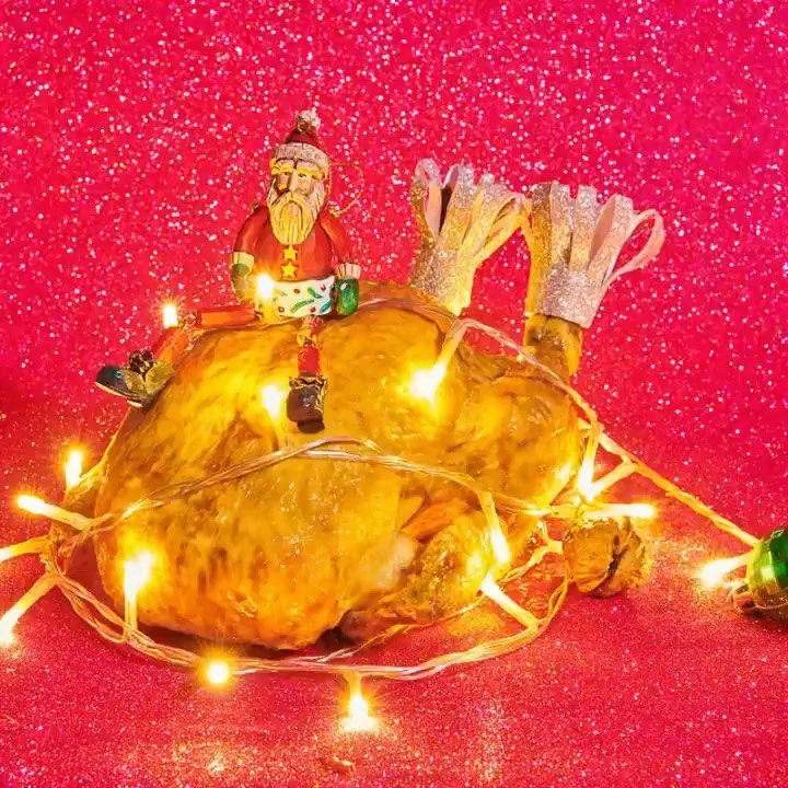 Holiday still life features a whole roasted turkey adorned with a Santa figurine, small gift props, festive string lights, and glittery antlers.