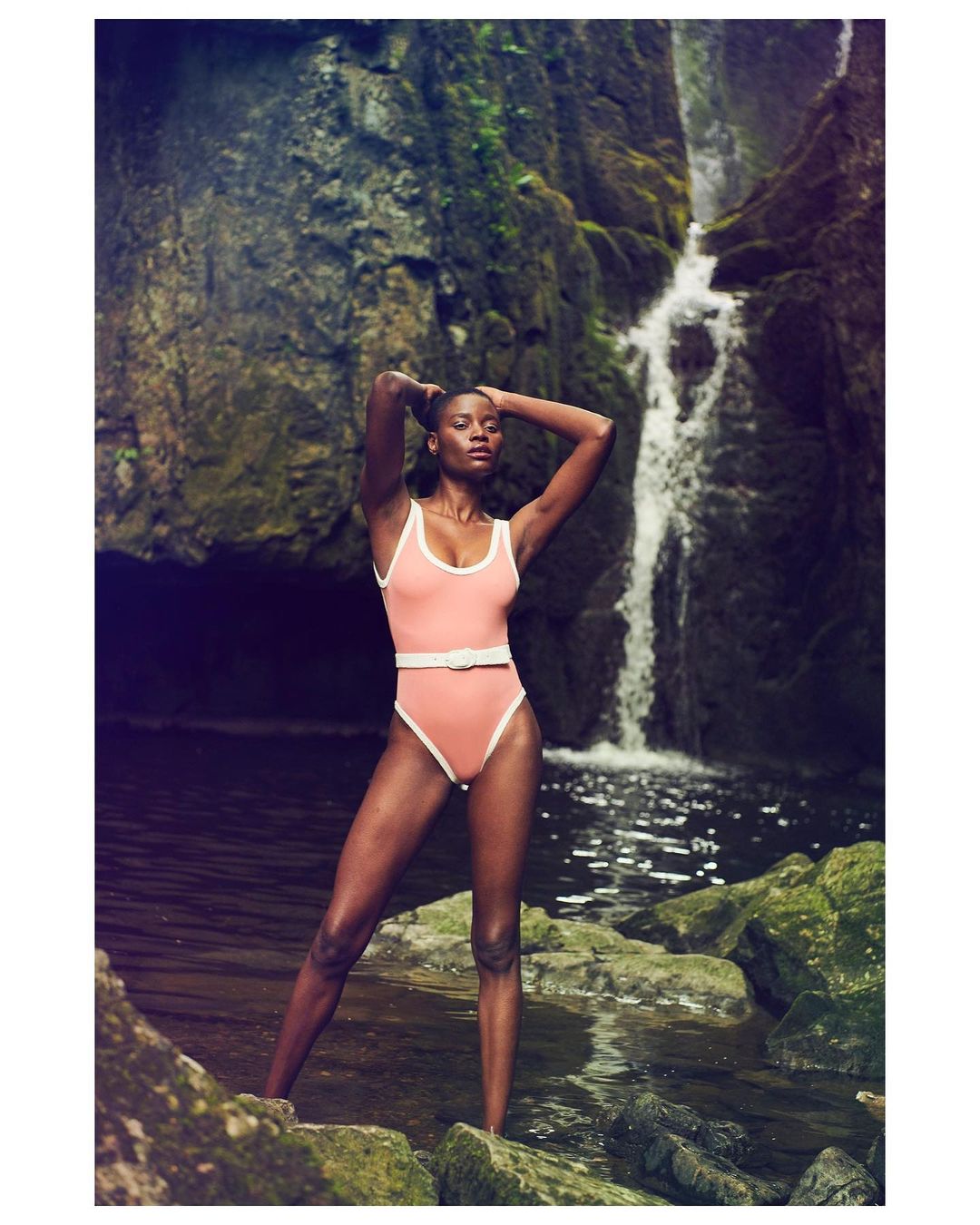 Woman in a pink swimsuit poses near a waterfall, surrounded by mossy rocks and lush greenery, capturing a serene and natural scene.