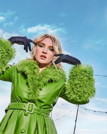 Woman wearing a bright green leather coat with furry collar and black gloves posing against a clear blue sky by Julia Kennedy