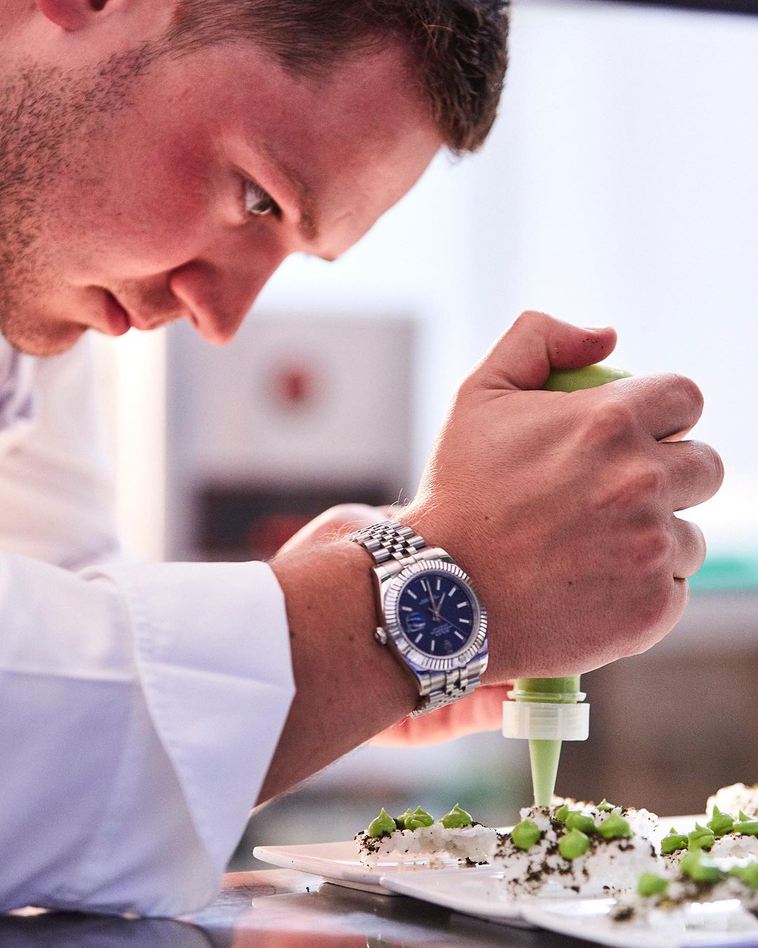 Adult male in chef's coat decorates dessert plate, wearing a wristwatch, indoor kitchen environment, food and beverage industry.