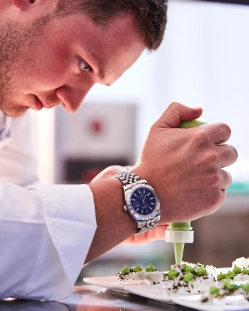 Adult male in chef's coat decorates dessert plate, wearing a wristwatch, indoor kitchen environment, food and beverage industry.