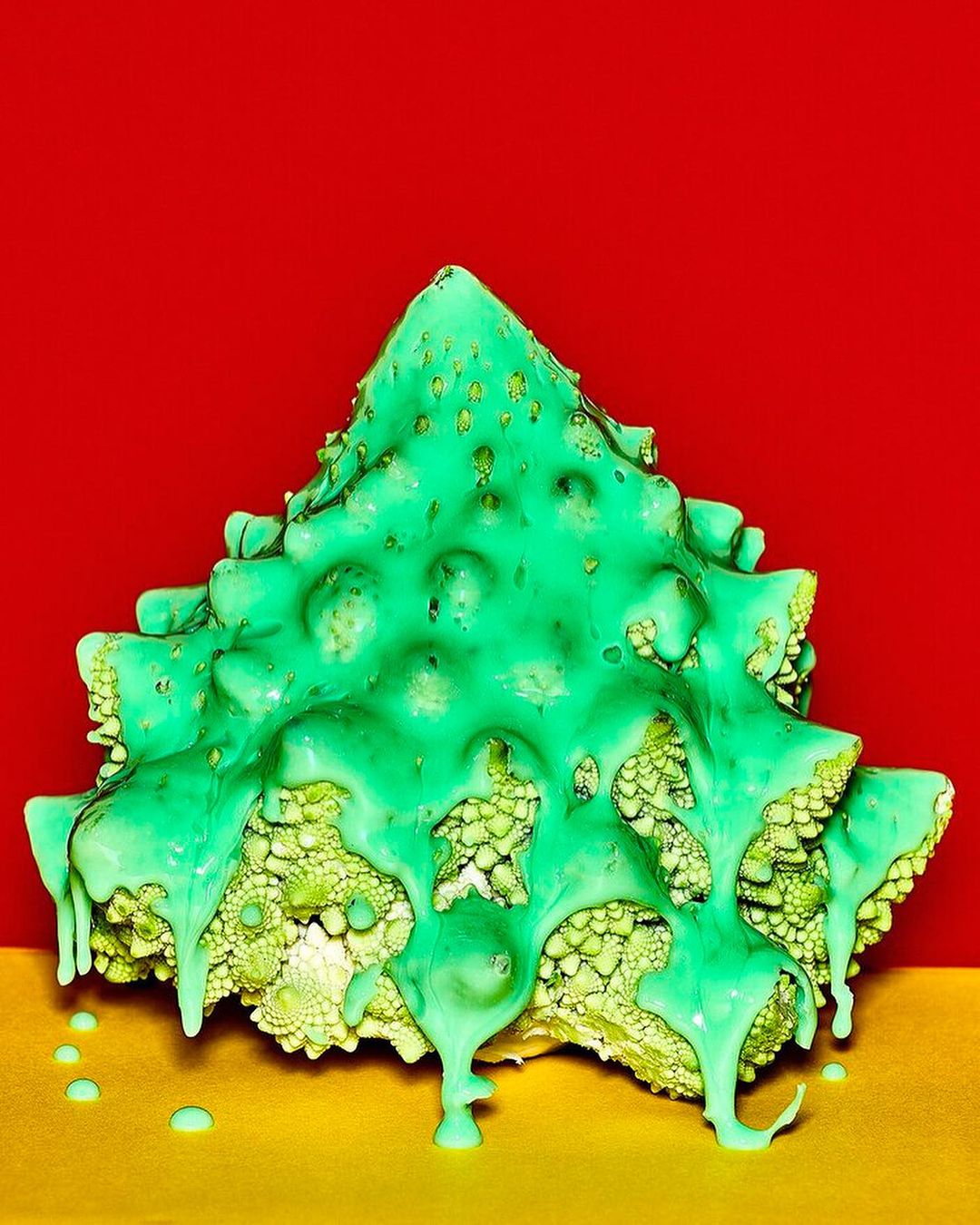 Abstract food photography featuring a Romanesco broccoli, styled as a Christmas tree, covered with dripping green sauce, set against a split red and yellow background.