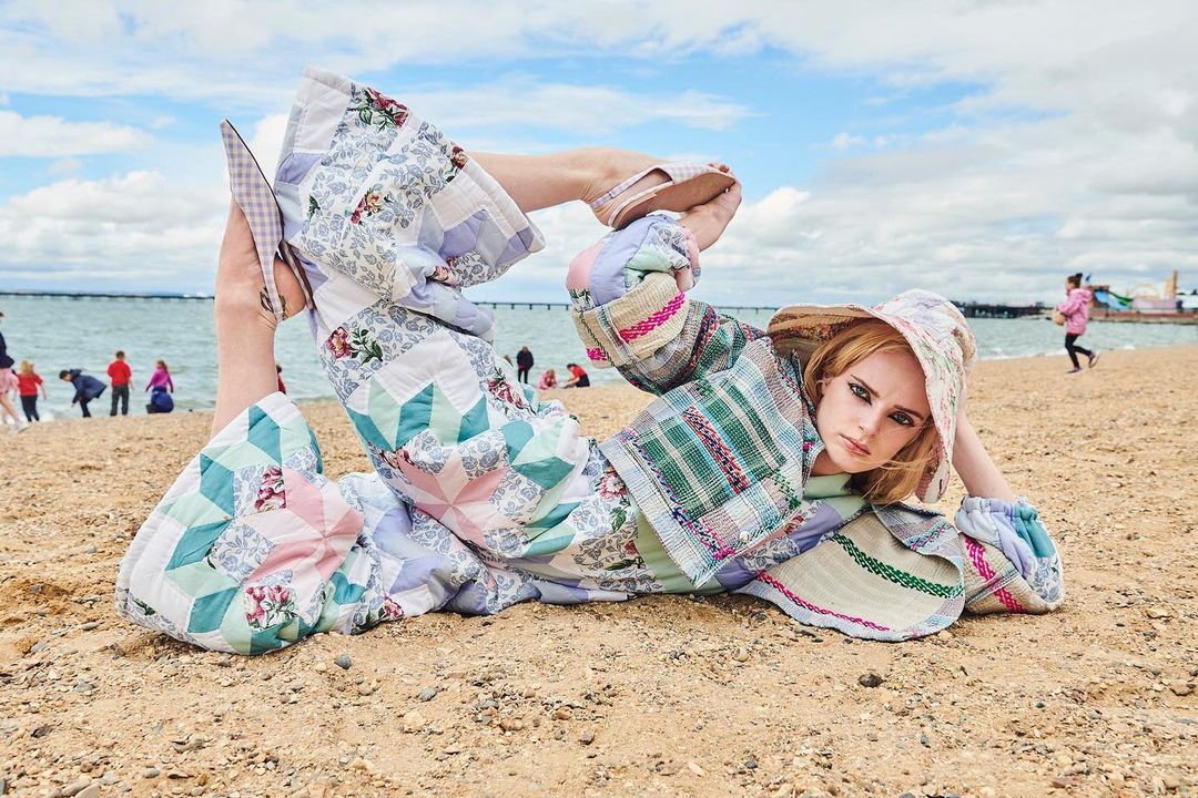 Model poses on a beach in a colorful patchwork outfit with ocean and people in the background on a partly cloudy day.