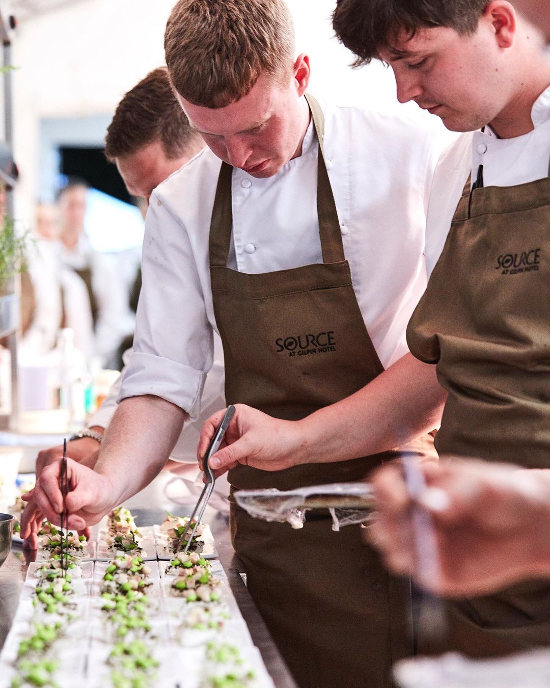 Chefs in aprons prepare small appetizers, using tweezers to carefully place toppings on the snacks at an indoor catering event.
