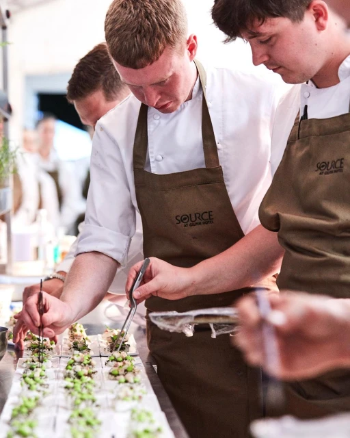Chefs in aprons prepare small appetizers, using tweezers to carefully place toppings on the snacks at an indoor catering event.