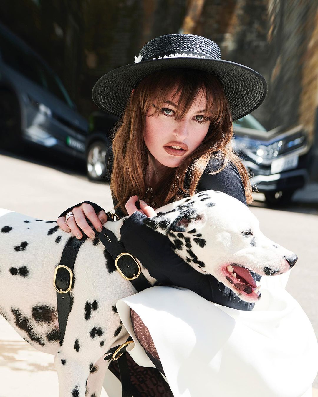 Adult woman in a hat, wearing black top and white skirt with a Dalmatian dog with a black leather harness outdoors on a sunny day.