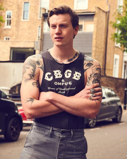 A cool and casual young adult man with arm tattoos, wearing a CBGB tank top, confidently poses outdoors against an urban street backdrop.