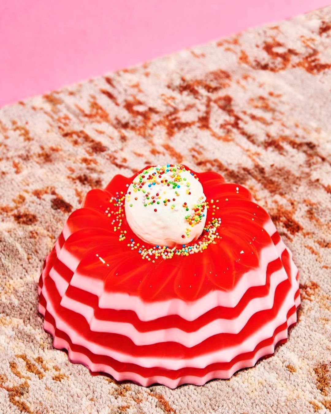 Close-up of a festive jello dessert with red and white stripes, topped with a scoop of white cream and sprinkles, on a patterned surface.