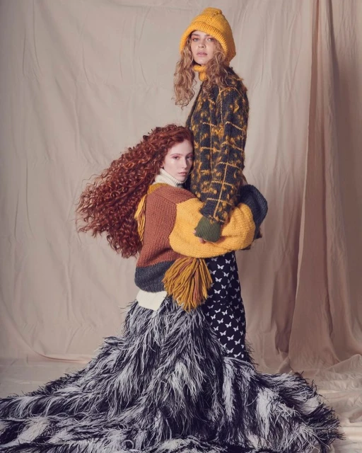 Two women in knitwear pose in a studio. One wears a yellow knit hat and patterned coat, while the other wears a sweater and feathered skirt.