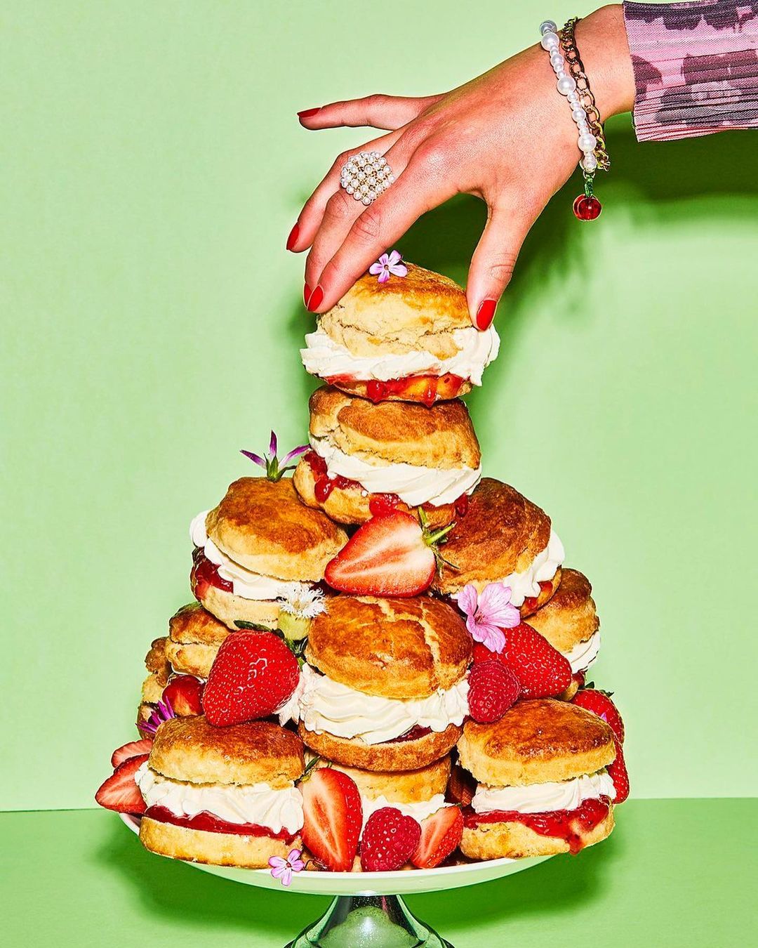 A woman's hand places the final scone on a tiered stack of scones, whipped cream, strawberries, and flowers against green backdrop.