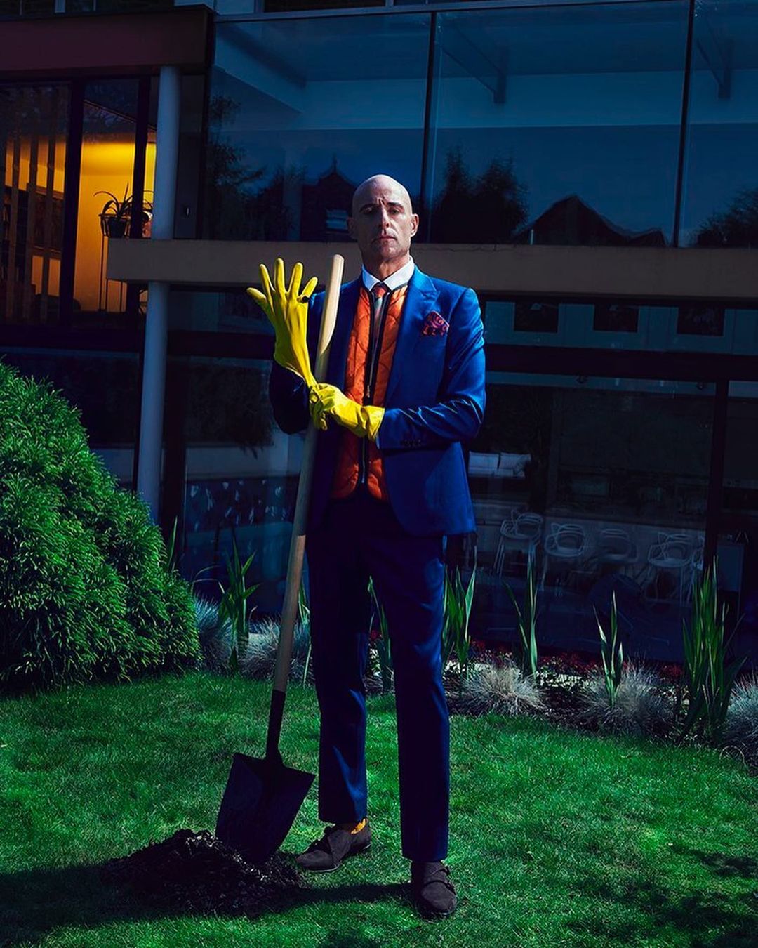 Well-dressed mature man in yellow gloves poses with shovel in garden with modern home at dusk. Concept shoot for real estate or lifestyle.