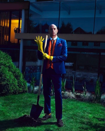 Well-dressed mature man in yellow gloves poses with shovel in garden with modern home at dusk. Concept shoot for real estate or lifestyle.